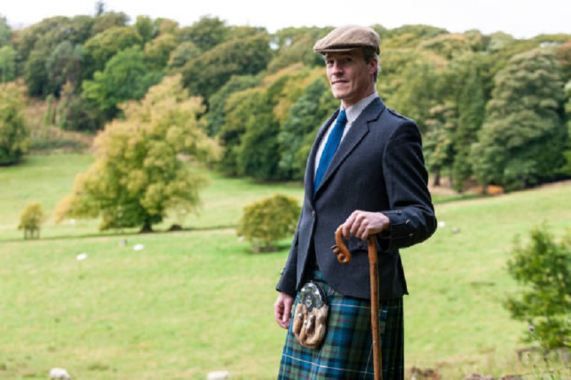 man wearing Scottish kilt and sporran standing on meadow leaning on cane looking at camera