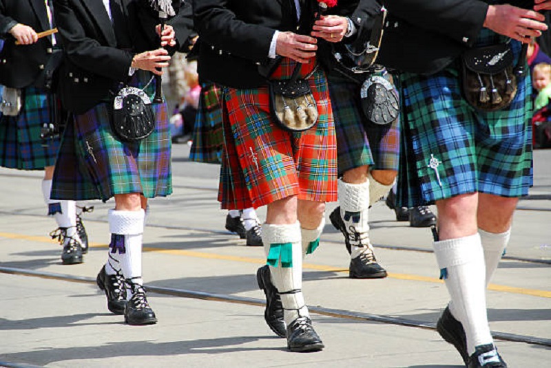 Captured from a low angle, this striking image showcases four individuals wearing kilts in various vibrant colors, marching with purpose. The kilts, reminiscent of both Scottish and Irish traditions, add a colorful and cultural flair to the scene. Witness the harmony of different hues as these individuals proudly showcase their heritage and the timeless appeal of kilts.